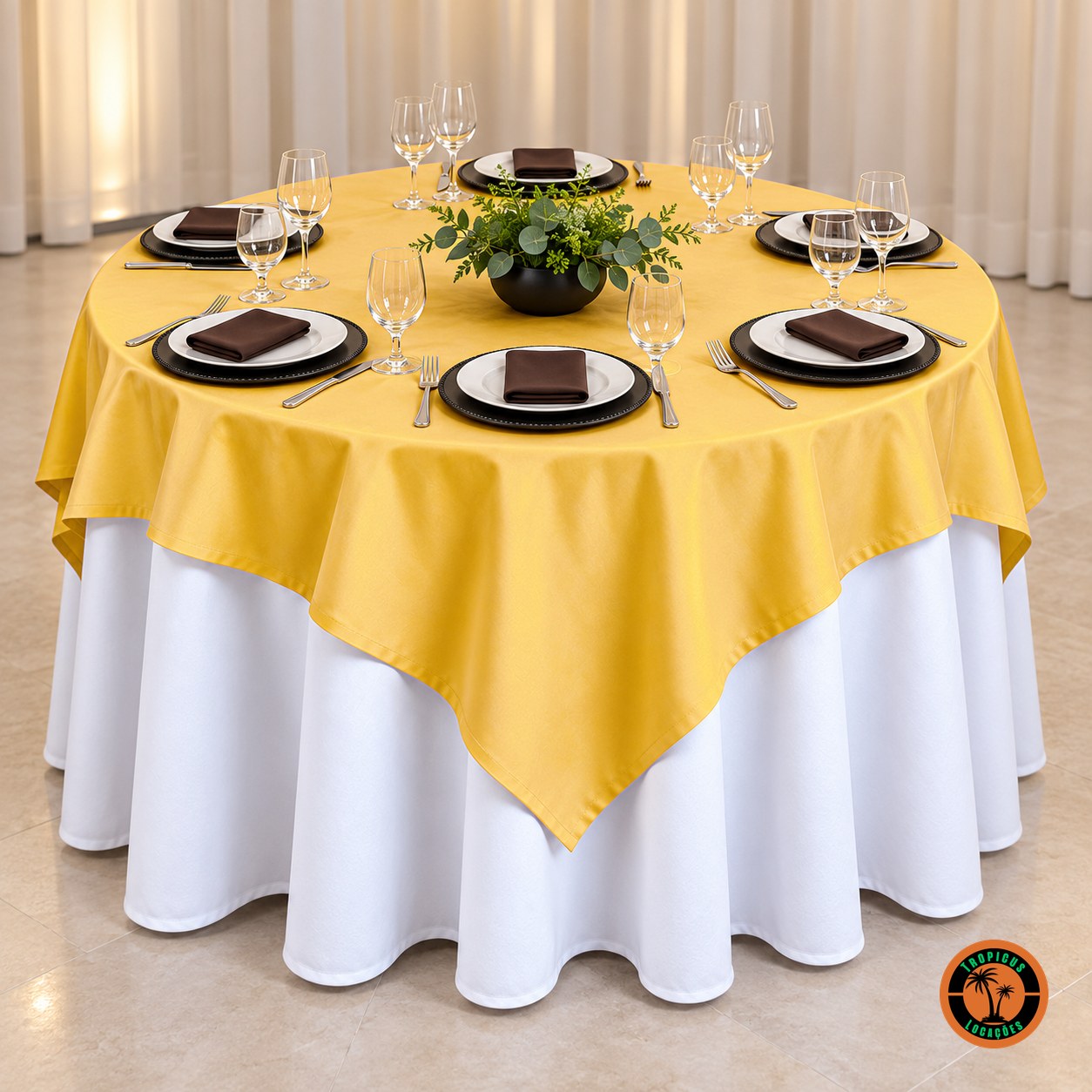 Mesa redonda com cobre-mancha amarelo-ouro sobre toalha branca, sousplats pretos e folhagens