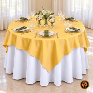 Mesa redonda com cobre-mancha amarelo-ouro sobre toalha branca, sousplats de vidro e flores brancas