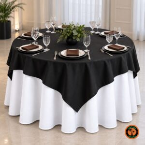 Mesa redonda com cobre-mancha preto sobre toalha branca, guardanapos marrons e folhagens