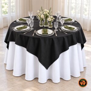 Mesa redonda com cobre-mancha preto sobre toalha branca, sousplats de vidro e flores brancas