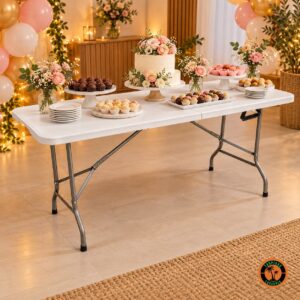 Mesa dobrável branca com bolo de aniversário, docinhos em boleiras, flores rosa e balões ao fundo